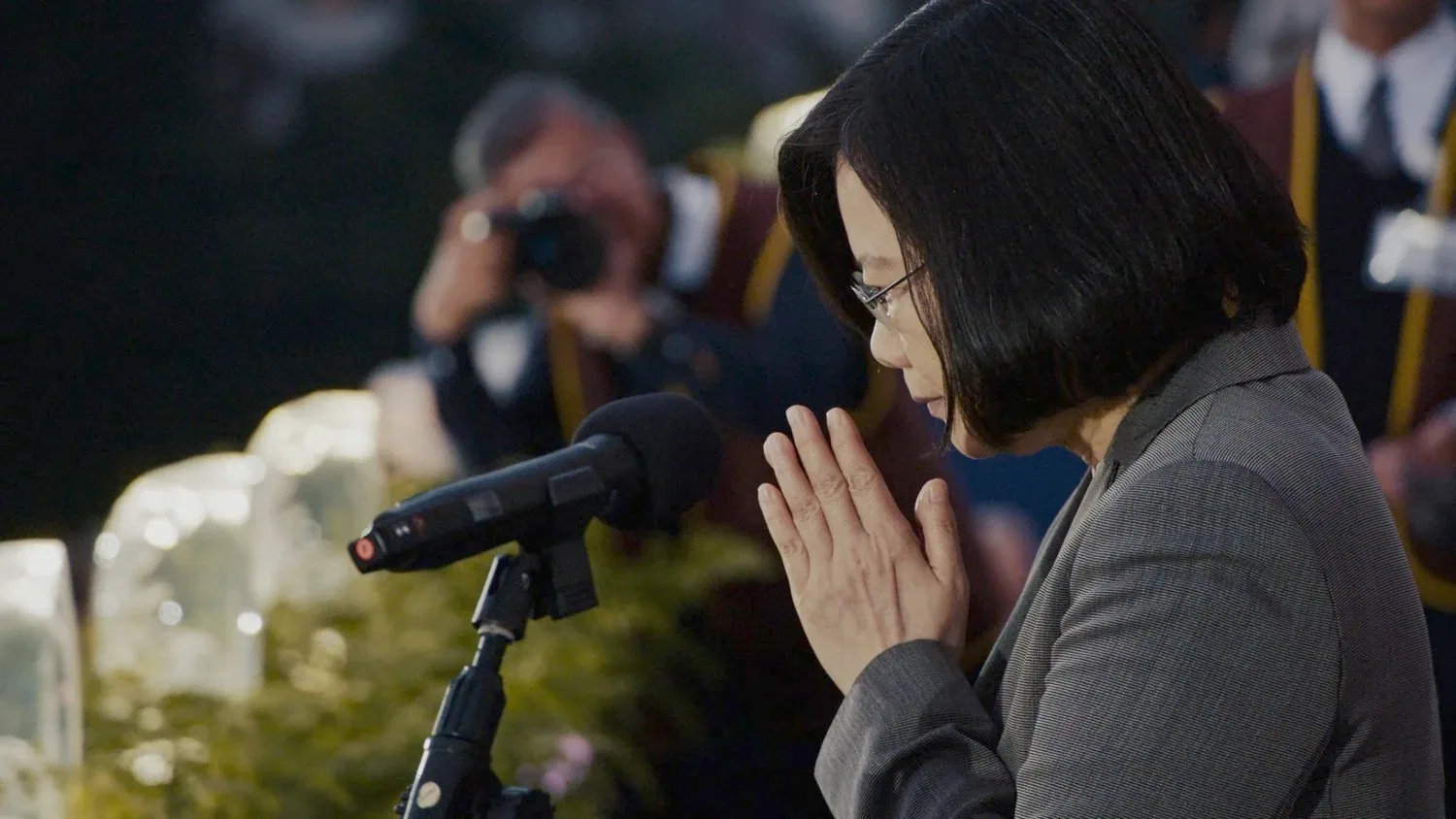Side close-up of Tsai Ing-wen with her head lowered and hands clasped, wearing glasses, a microphone in front of her, and a photographer in the background raising a camera to photograph her