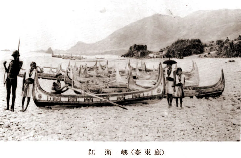 約 1931 年的紅頭嶼（蘭嶼舊稱）海灘上，一艘大型拼板舟停泊在卵石灘上，艏艉高翹、側板刻滿雕紋，幾位穿著傳統服飾、頭戴遮陽圓帽的達悟族人立於舟旁，背景是島上的山林與海灣