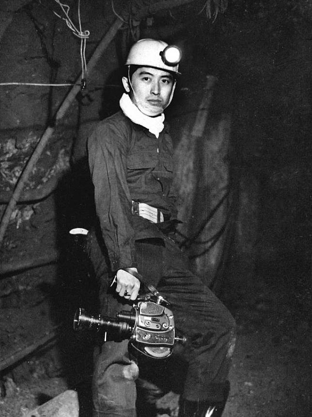 El joven Zhang Zhaotang viste un uniforme de trabajo y un casco de seguridad de minero con linterna frontal, de pie en un túnel oscuro, sosteniendo una cámara de cine de 16 mm en su mano derecha