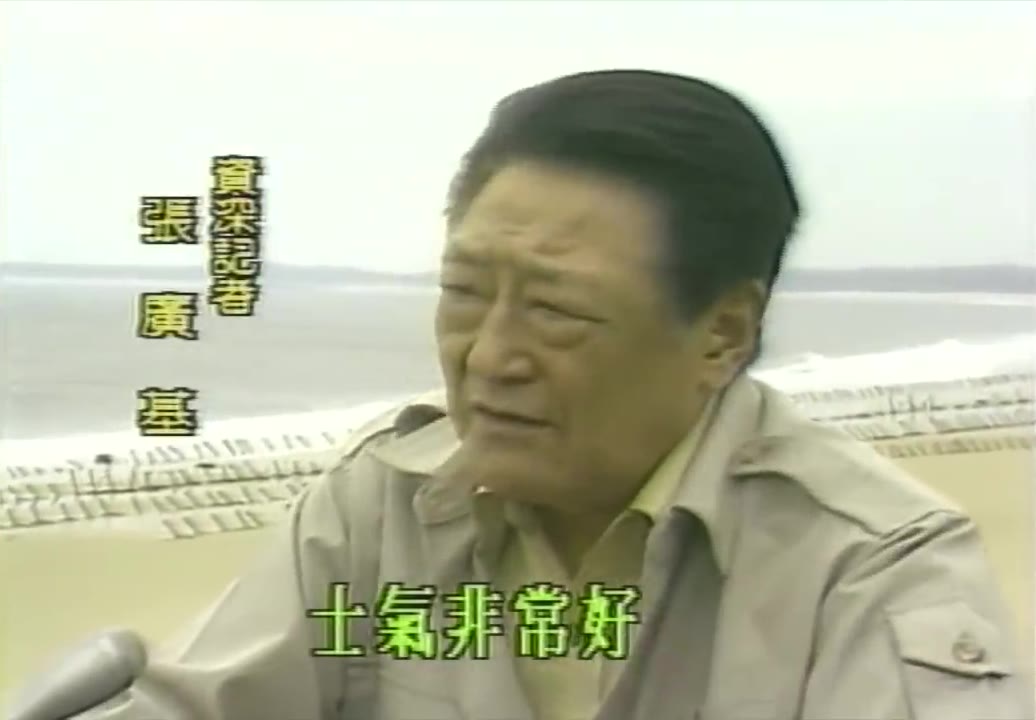 El veterano reportero Zhang Guangji de pie en la playa de Kinmen durante una entrevista, vistiendo un uniforme de caza color caqui, con olas y una fila de obstáculos antidescenso al fondo, con una tarjeta de texto en vertical a la izquierda que dice «Veterano reportero Zhang Guangji»