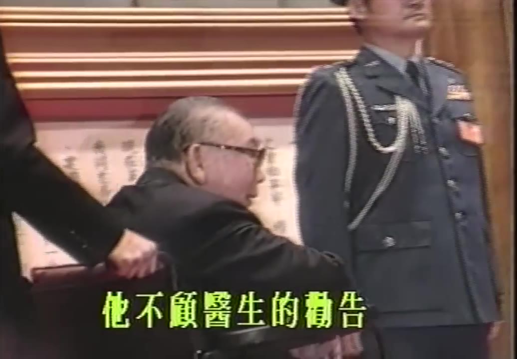 El anciano Chiang Ching-kuo, con gafas, sentado en una silla de ruedas, siendo empujado hacia el salón, con un oficial uniformado de pie a la derecha, y un subtítulo abajo que dice «Ignoró los consejos del médico»