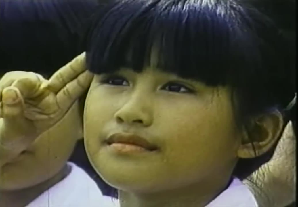 Primer plano de una niña pequeña con sombrero y flequillo recto levantando la mano en saludo, con expresión concentrada