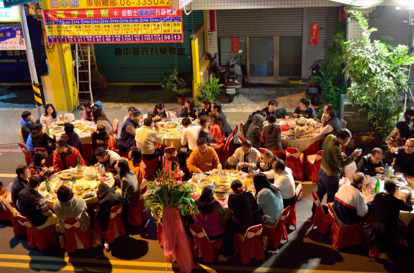 Round tables with red chair covers in Tainan at night, one after another, filled with guests, with storefronts with rolling shutters down and motorcycles parked on the roadside in the background