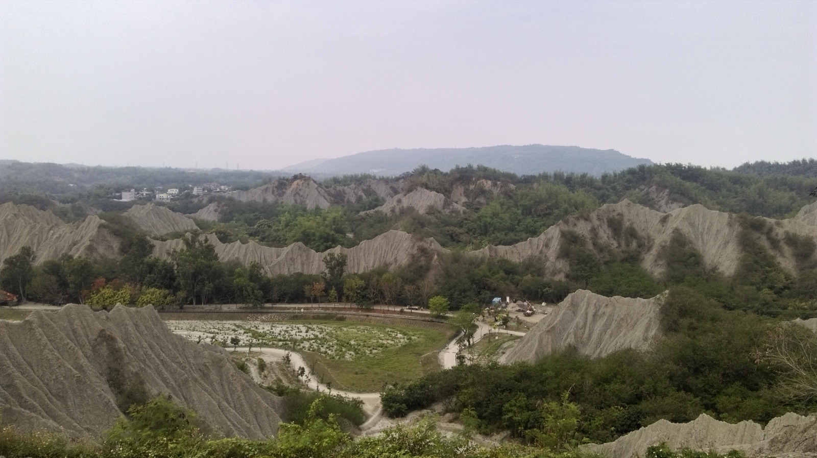 The mudstone badlands of the Gutingkeng Formation in Kaohsiung, with gray-white ridges exposed one after another, almost barren, with sparse green trees and small settlements in the distance