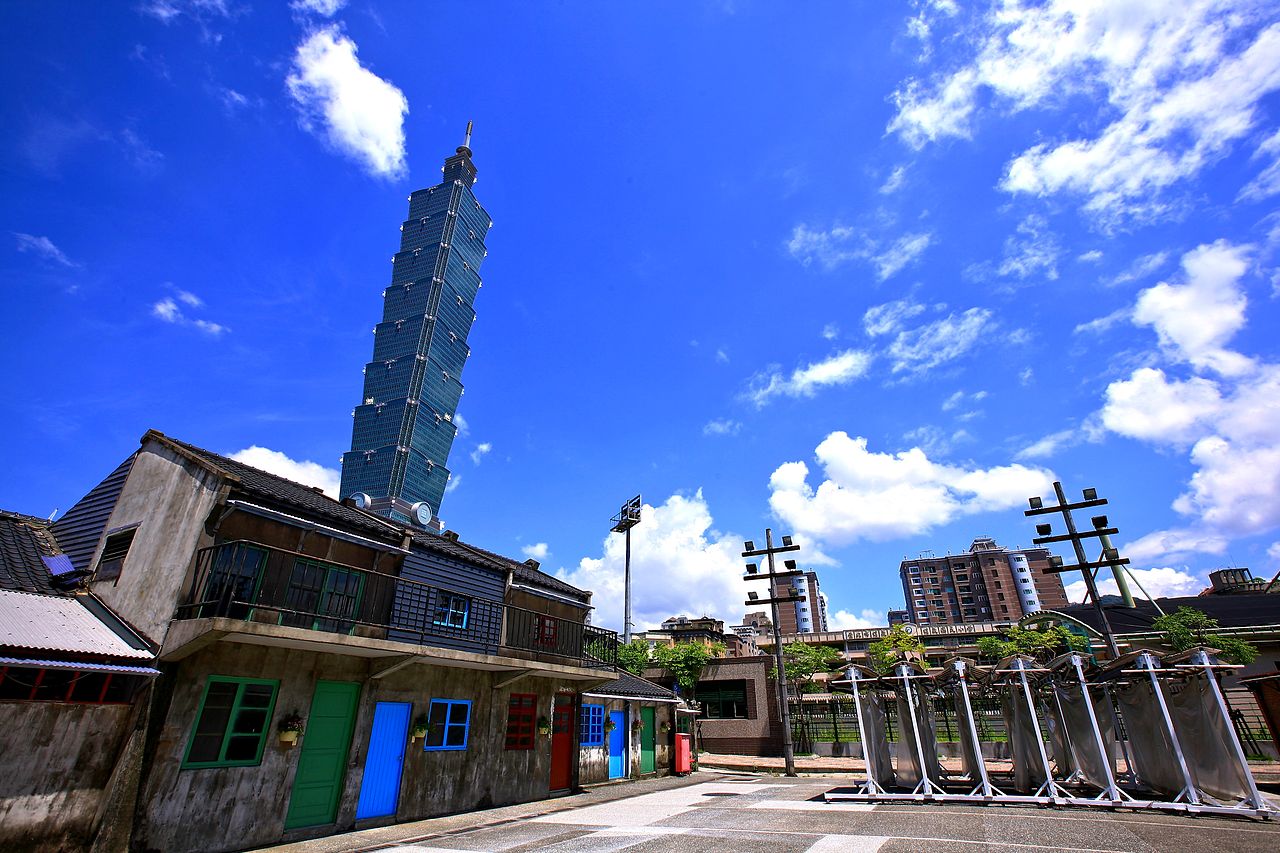 四四南村全景。保留下來的 4 棟磚瓦平房圍出一塊草地廣場,背景是 2013 年正在建造的信義計畫區高樓。1948 年的眷舍跟 21 世紀的鋼骨玻璃並列。