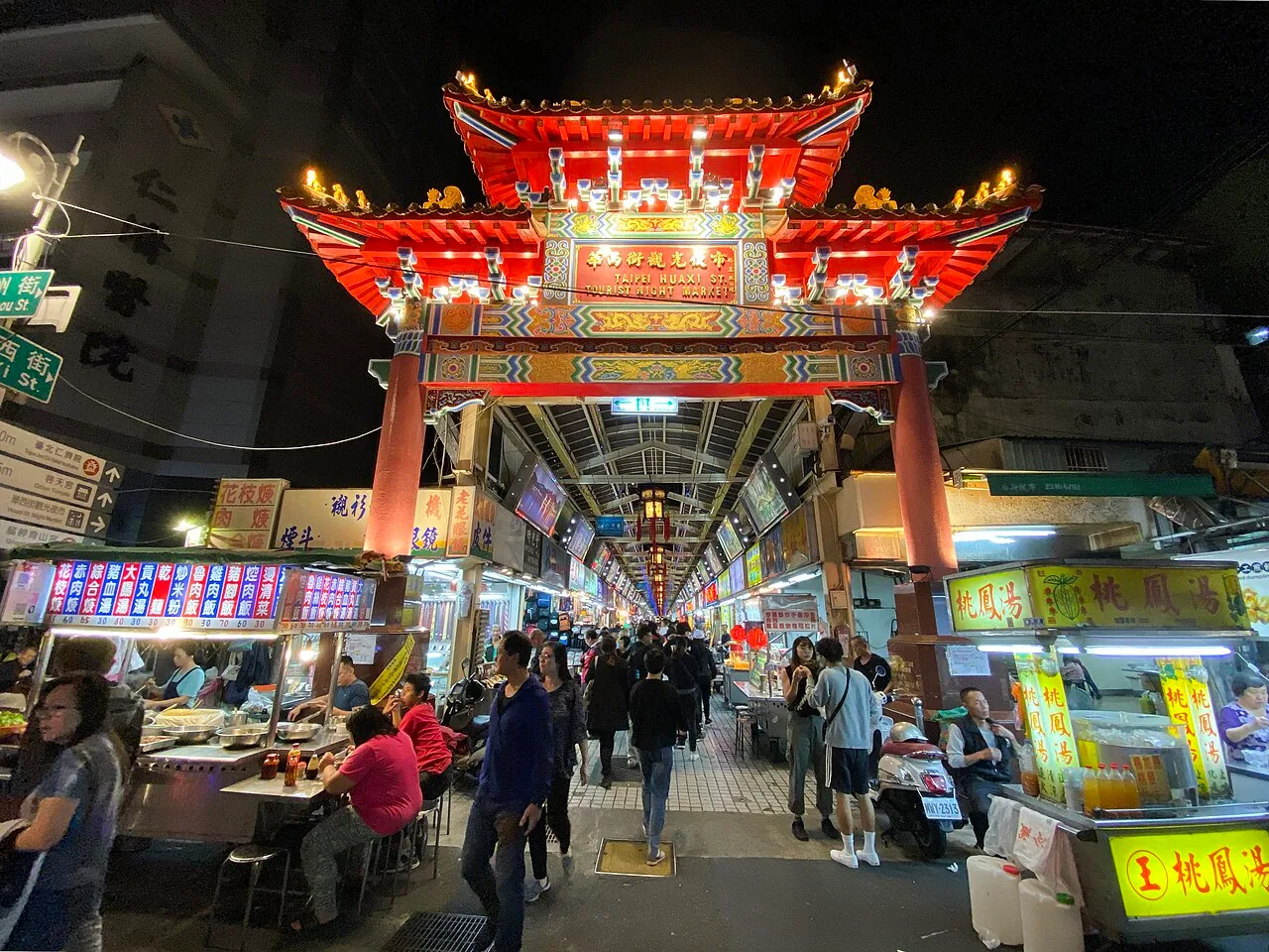 Arcada del mercado nocturno turístico de la calle Huaxi. En 1972, los burdeles públicos fueron concentrados en Baodouli, actual zona de la calle Huaxi. Tras la abolición de la prostitución en 1997, se transformó en un mercado nocturno turístico centrado en los bocadillos; los letreros de las tiendas de serpiente son su residuo histórico más visible.