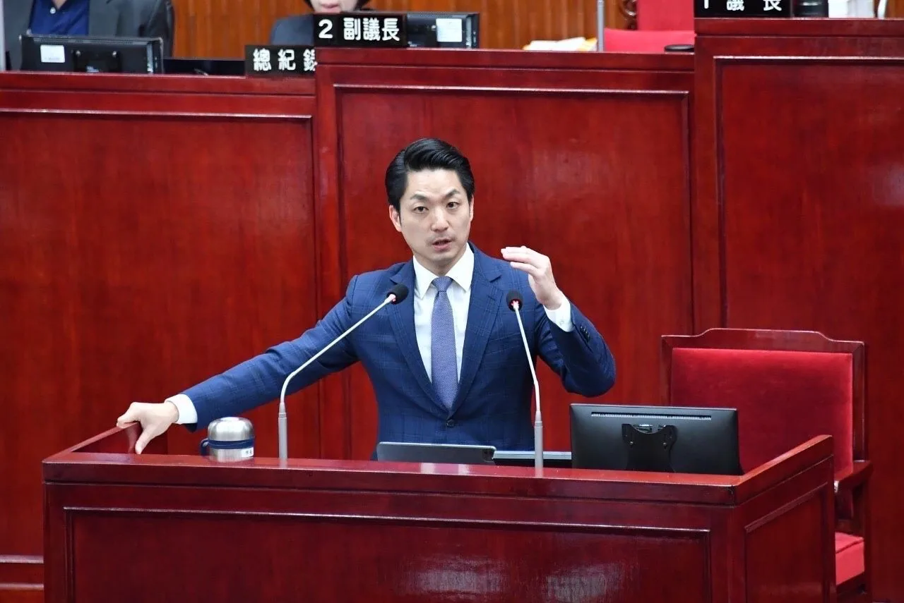 Chiang Wan-an steht vor dem Befragungstisch des Stadtrats von Taipeh, eine Hand stützt sich auf die Tischplatte, während er mit der anderen Hand Gesten macht, um Fragen zu beantworten; hinter ihm befindet sich der Sitz des Ratspräsidenten.