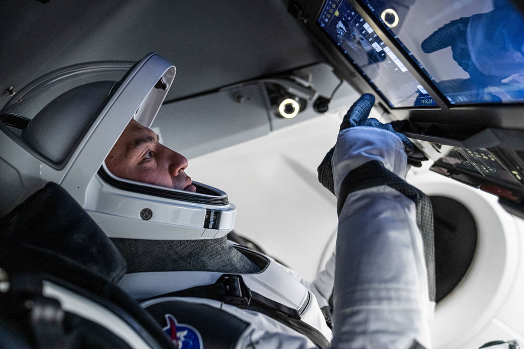 Lindgren in a blue NASA jumpsuit, arms crossed, standing in front of a Crew Dragon mockup capsule at SpaceX headquarters in Hawthorne, California. His NASA patch and American flag are clearly visible on his shoulders