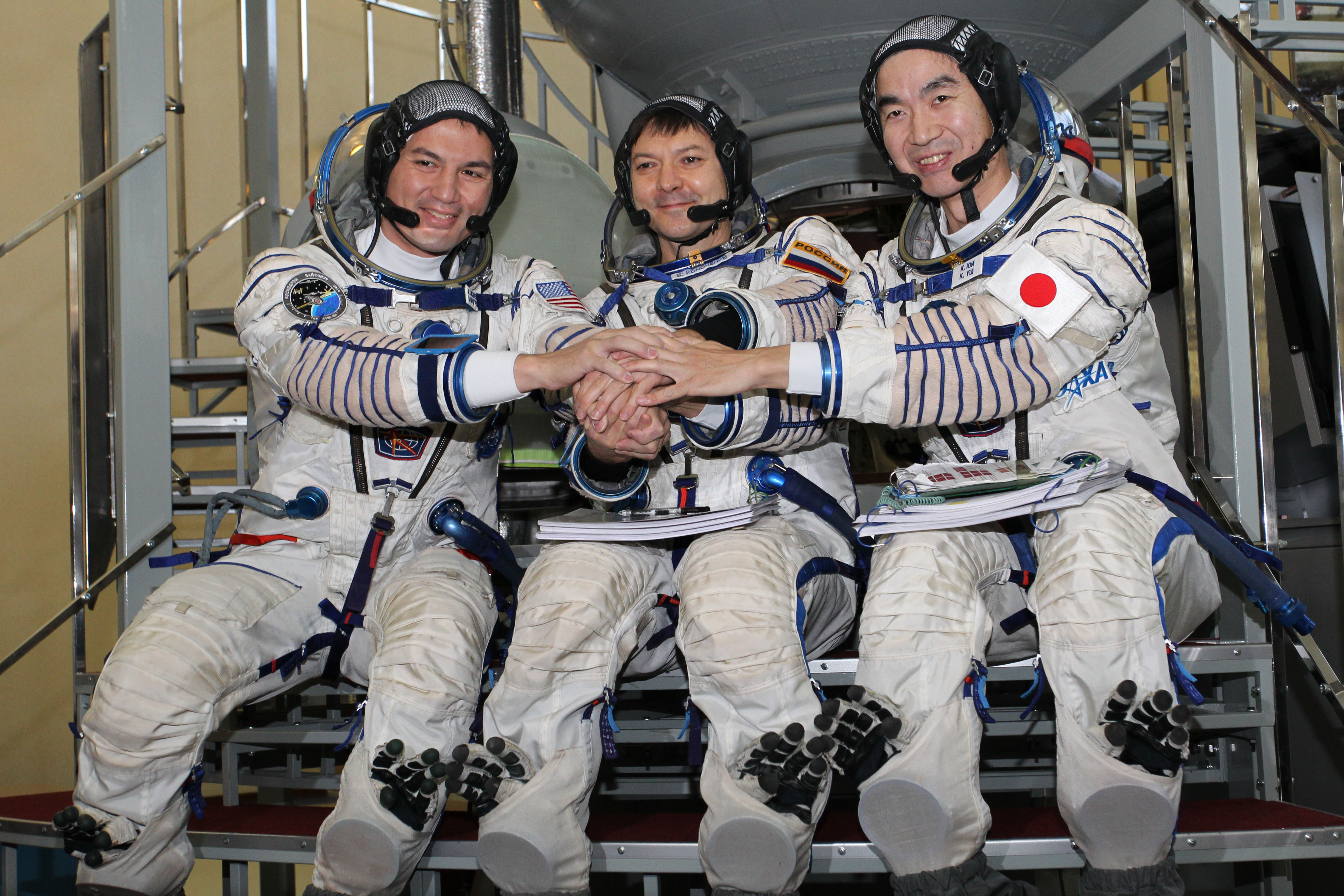 Three astronauts standing in front of the Soyuz TMA spacecraft mock-up at the Gagarin Cosmonaut Training Center in Star City, Russia. From left: Lindgren (NASA), Oleg Kononenko (Russia), Kimiya Yui (Japan), all wearing blue Expedition 42/43 backup crew jumpsuits