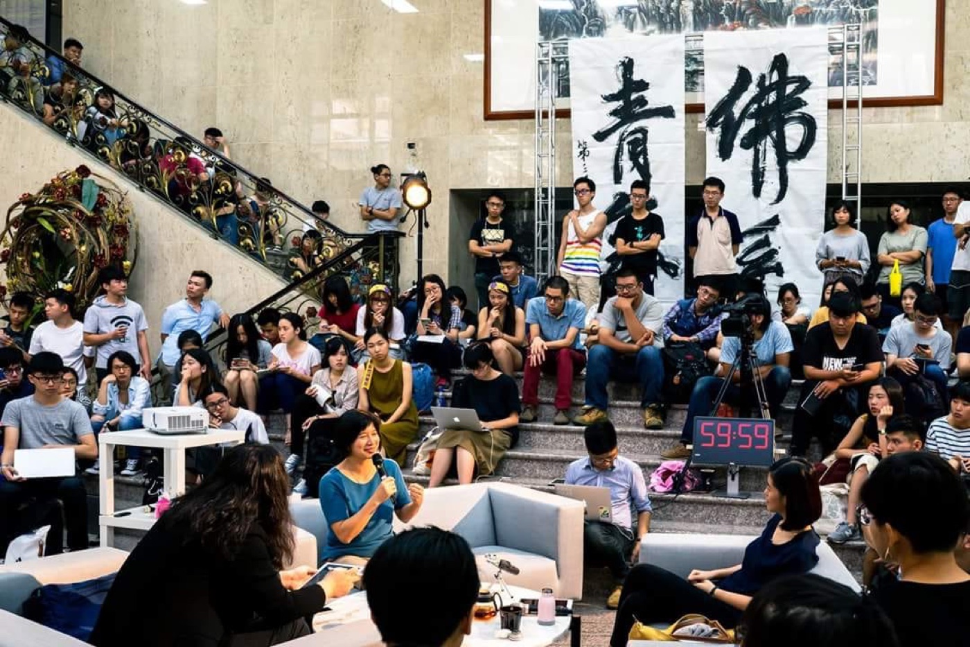 一名講者站在台上手持麥克風,後方掛著「佛系青年」的書法布條,旁邊有一個顯示 59:59 的倒數計時鐘