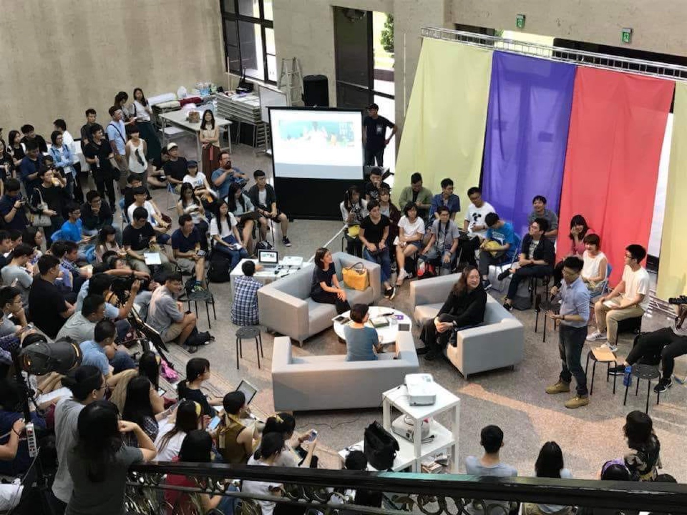俯瞰視角:三張沙發排成一個三角形,三名講者朝內而坐、背向四周環繞的觀眾,沙發後方分別垂下紅、藍、黃三色布幕