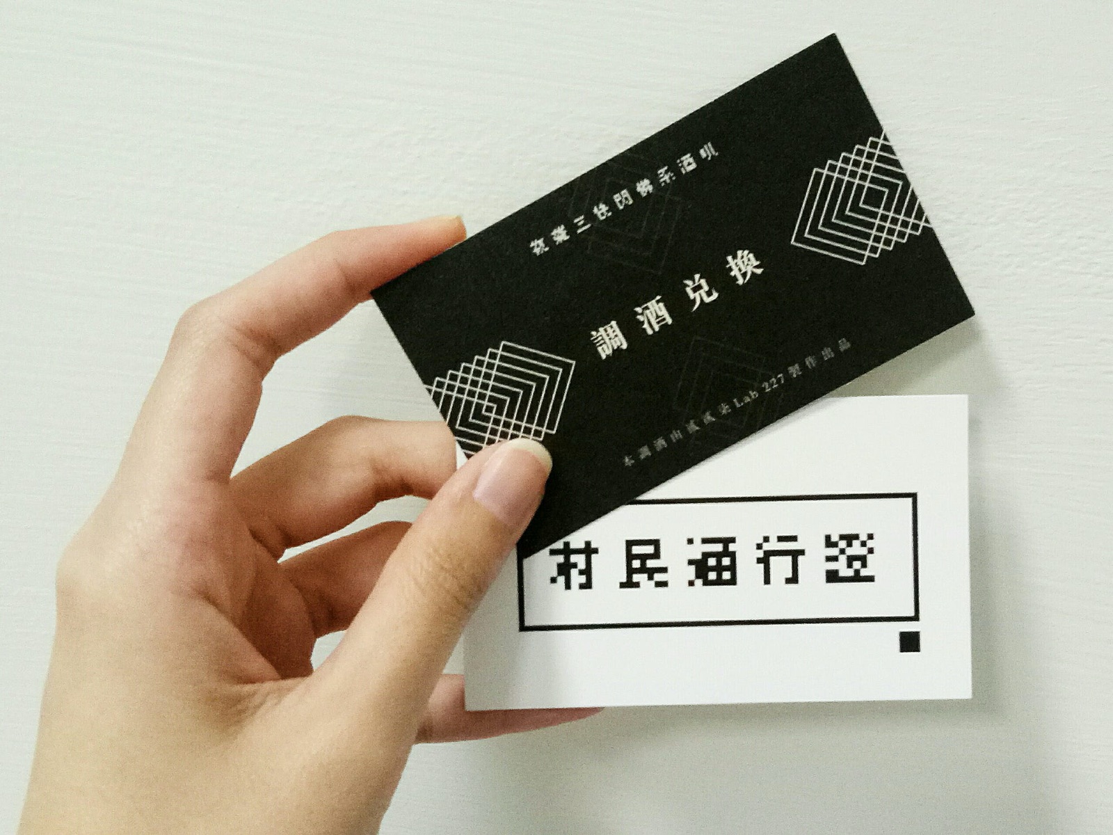 一隻手拿著兩張券,一張印著「村民通行證」、一張印著「調酒兌換」,券面用幾何感很強的點陣字體設計