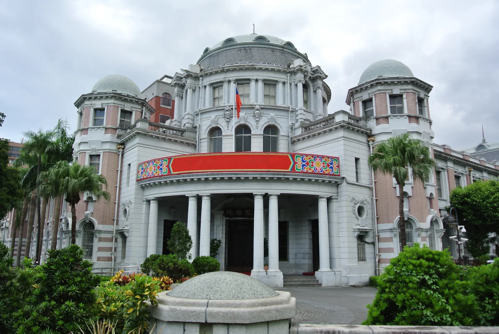 監察院正面，日治時期落成的建築有一大兩小三個圓頂，白色列柱門廊上方掛著紅底彩繪橫幅，門楣刻著監察院三字，兩側種著椰子樹，天空陰雲密布
