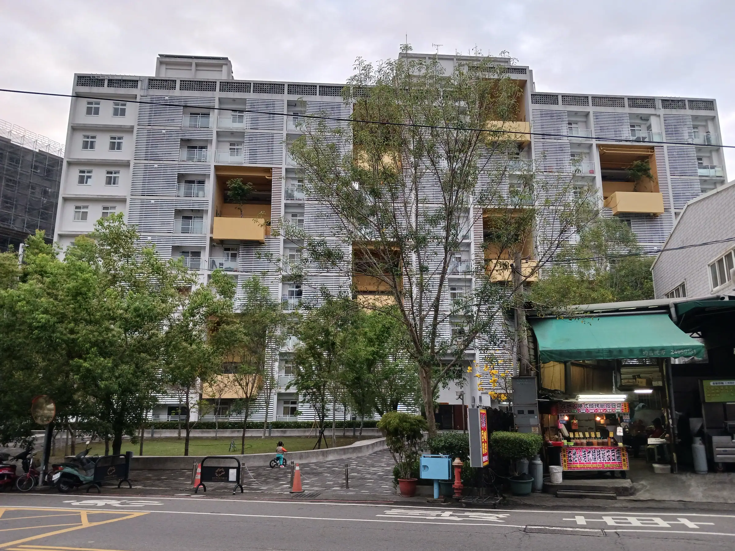光正社會住宅的臨街立面:陽台外掛遮陽格柵,樓下是街邊綠地與騎樓小吃攤,孩子在草地上騎滑步車