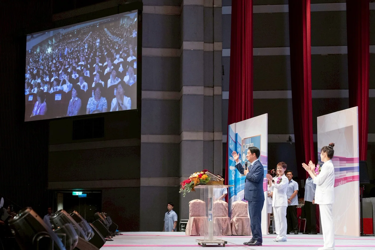 Presidente Lai Qing-de acenando ao lado do púlpito, com duas enfermeiras de uniforme branco e broches de peito aplaudindo em cada lado, tela grande ao fundo projetando enfermeiros lotando o auditório, Grande Conferência Conjunta de Celebração do 115º Dia Internacional da Enfermeira