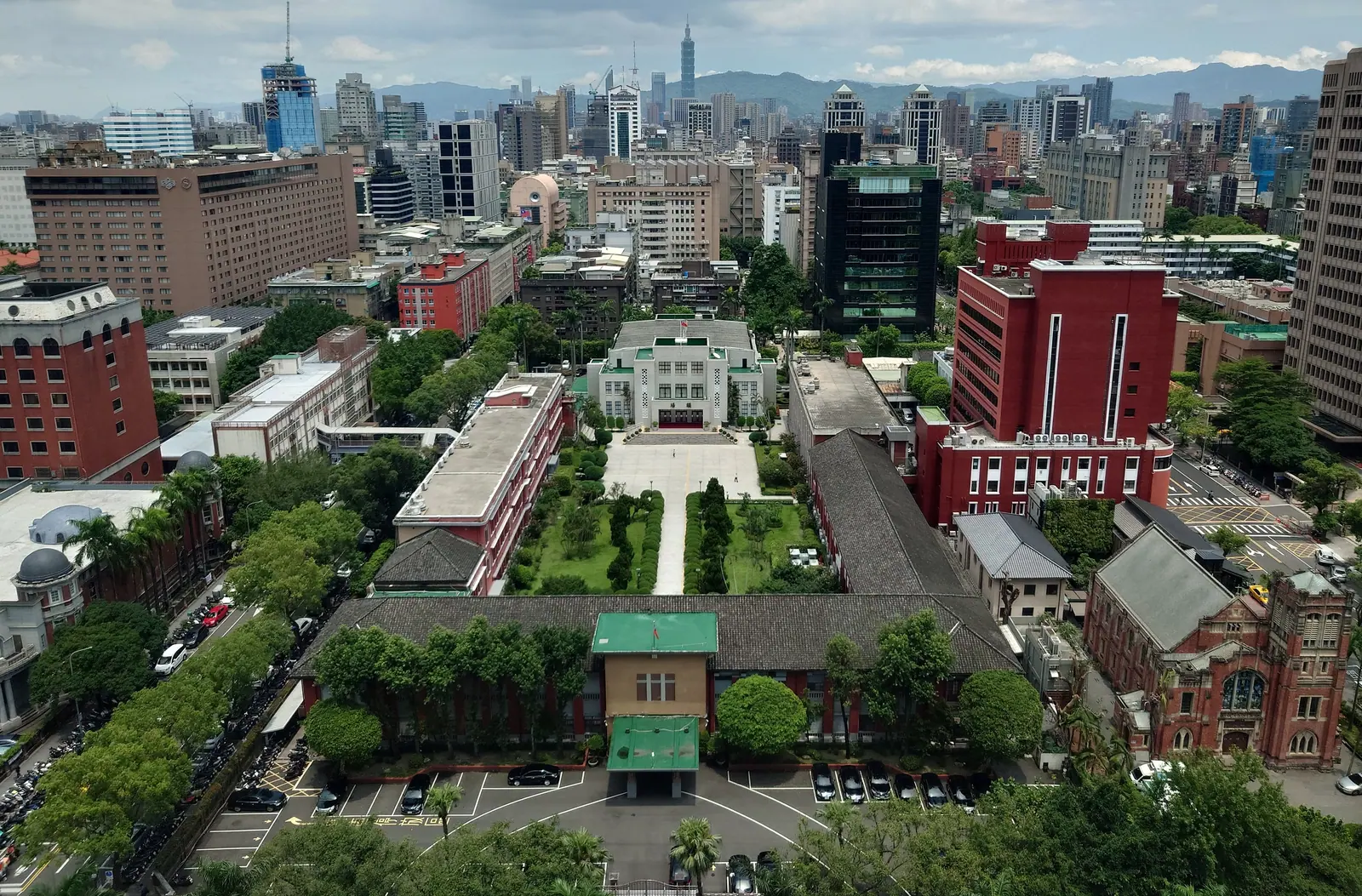 從高處俯瞰立法院院區，綠色屋頂的低矮議場與辦公樓圍出中央庭園與修剪整齊的樹列，四周是密集的台北市街景，遠方可見台北 101 與環繞的山稜