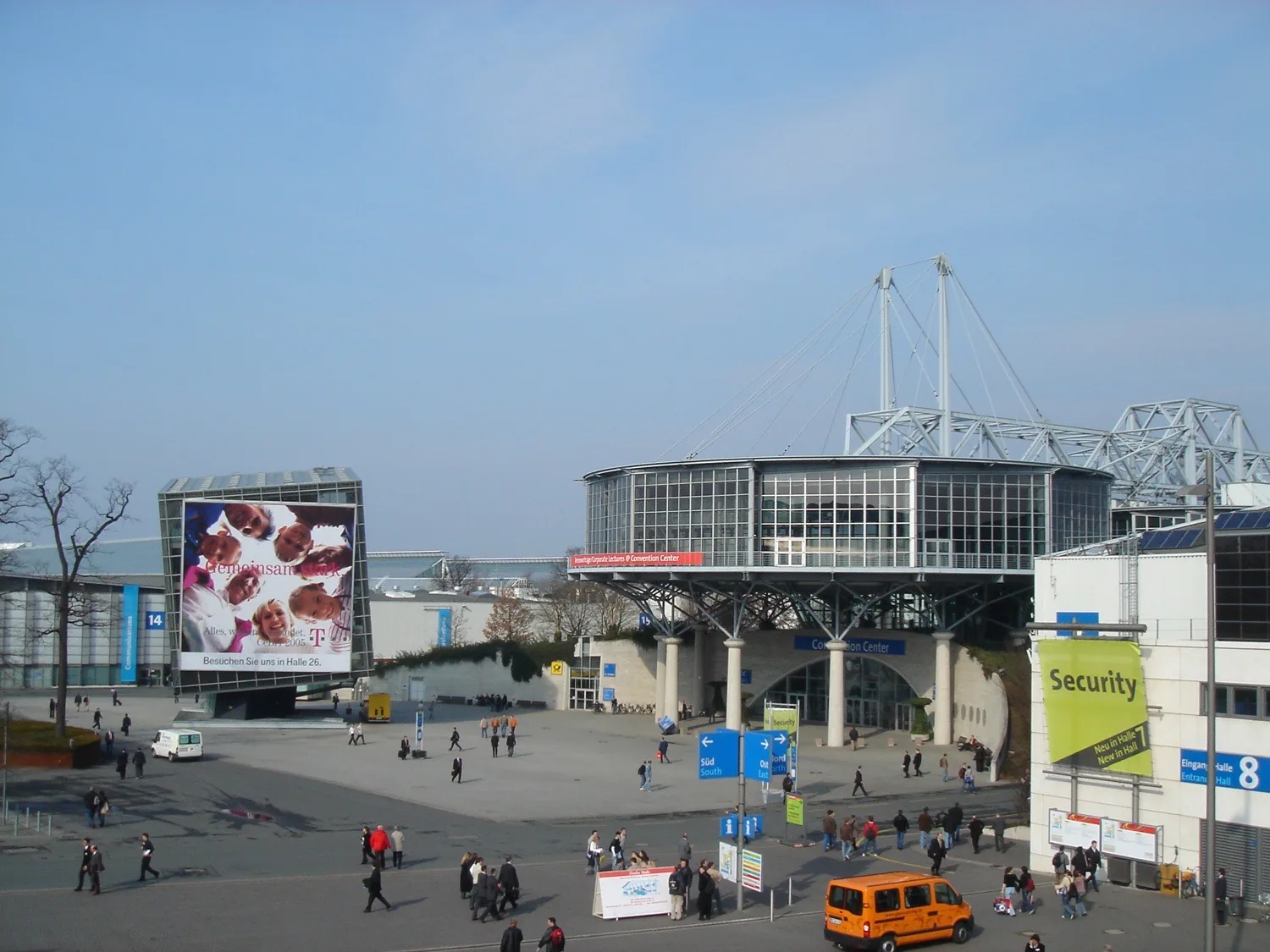 El recinto de CeBIT 2005 en Hannover, Alemania, lleno de visitantes; a ambos lados de los amplios pasillos se alinean puestos de empresas tecnológicas, con techos altos y abundantes letreros, mostrando la escala que tenía entonces la mayor feria informática del mundo