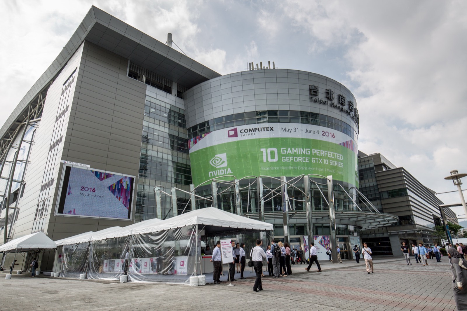 南港展覽館一館外觀，玻璃帷幕建築上高掛 COMPUTEX TAIPEI 大型展覽看板，門口聚集準備入場、掛著識別證的參觀人潮，這裡是台北國際電腦展近年的主場館