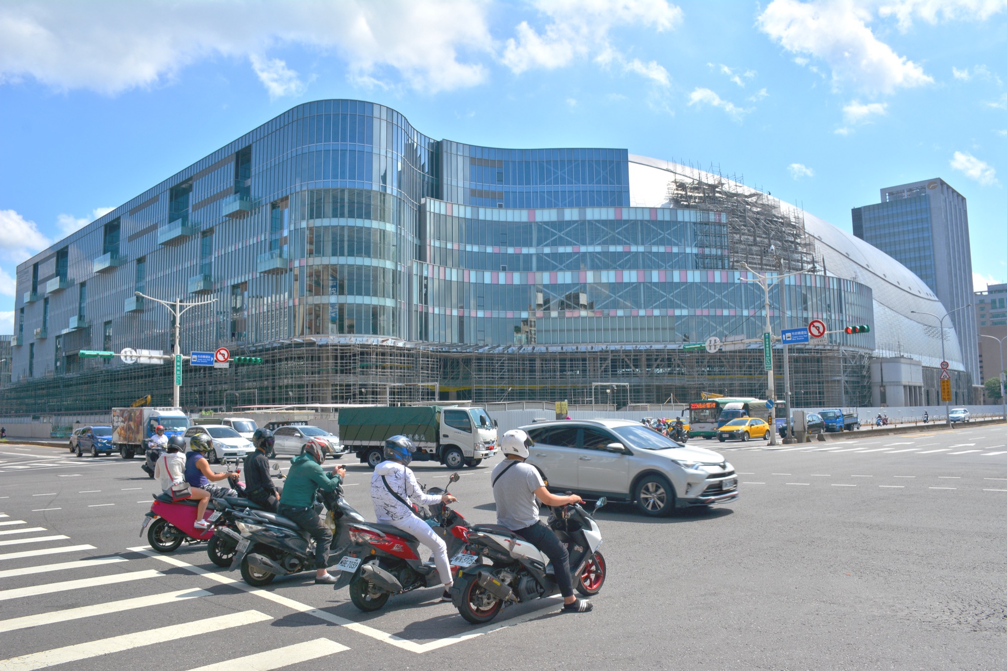 2020 年 6 月 21 日上午台北大巨蛋施工現場街景,遠景大巨蛋鋼構鐵皮外殼仍在搭建中,前景一輛 Hino 300 卡車正經過忠孝東路靠近國父紀念館捷運站 5 號出口的斑馬線,反映台北最大型體育場館長達十多年的施工現實與大林組在這個 6.5 萬噸圓形鋼管巨蛋的施工管理角色